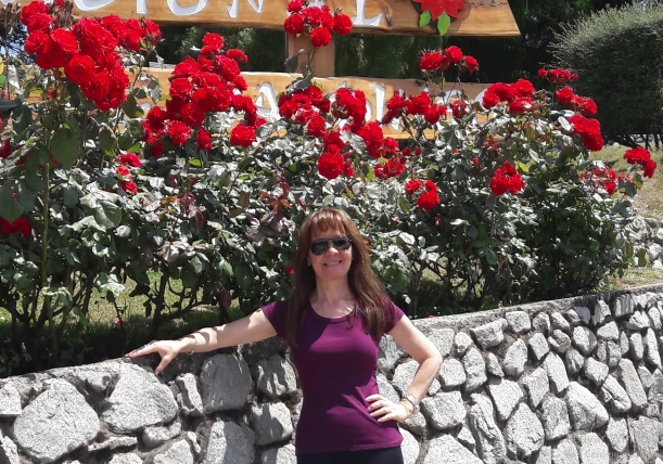 Imagen sonriente de la consultora, parada junto a un muro de piedras junto a rosales con flores rojas